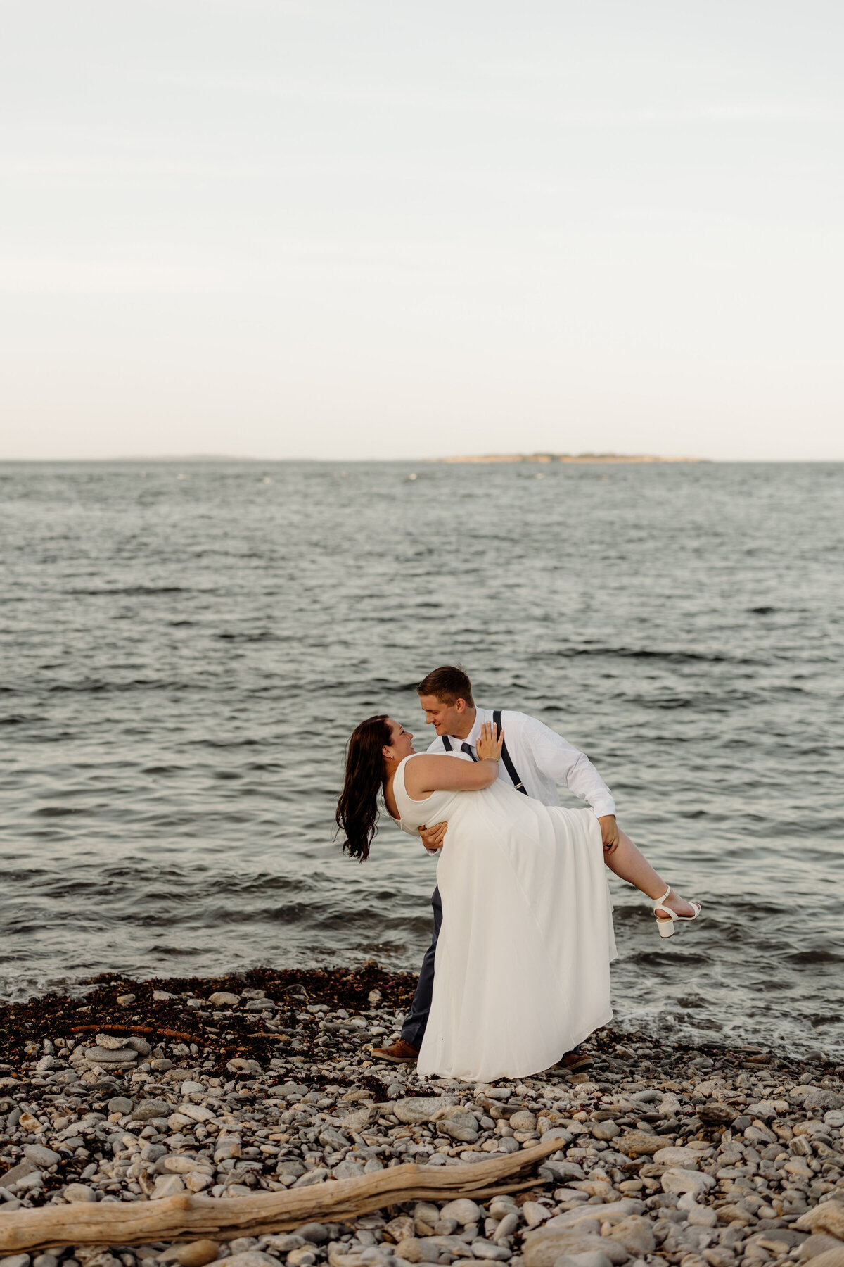 A couple is on a rocky beach by the sea, captured in the candid style of an Elopement Photography New England session. The man, in a white shirt and suspenders, playfully lifts the woman in a white dress. Their joyful smiles mirror the calm waters and distant island backdrop.