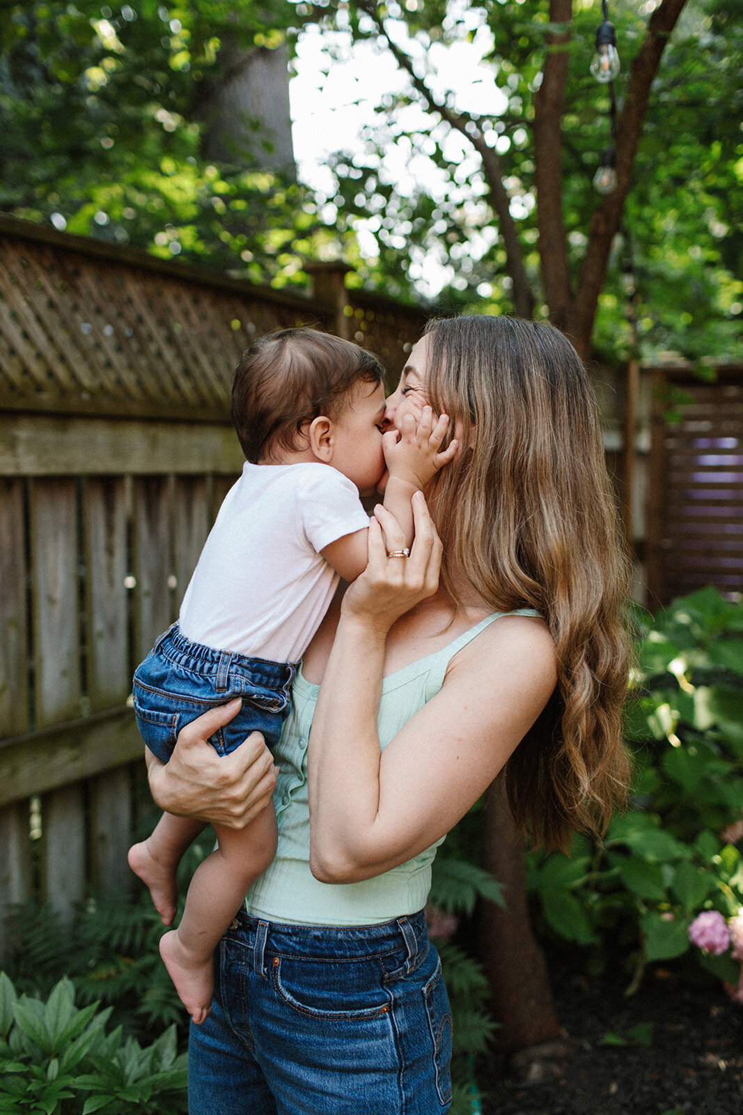 A woman holds a baby in a garden, both grinning as the little one playfully touches her face. With a wooden fence and lush greenery as backdrop, this charming moment captured by a Toronto family photographer highlights pure joy.