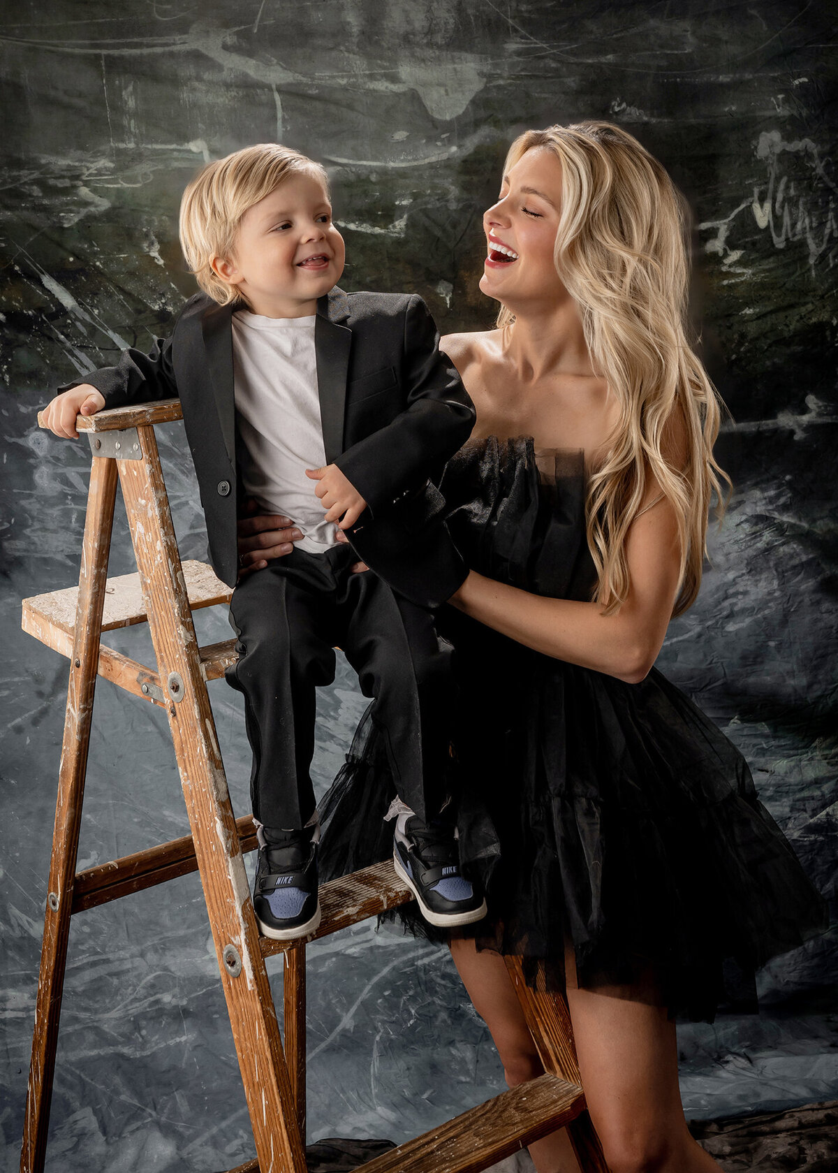 A boy and his mom look at each other laughing while he stands on a ladder