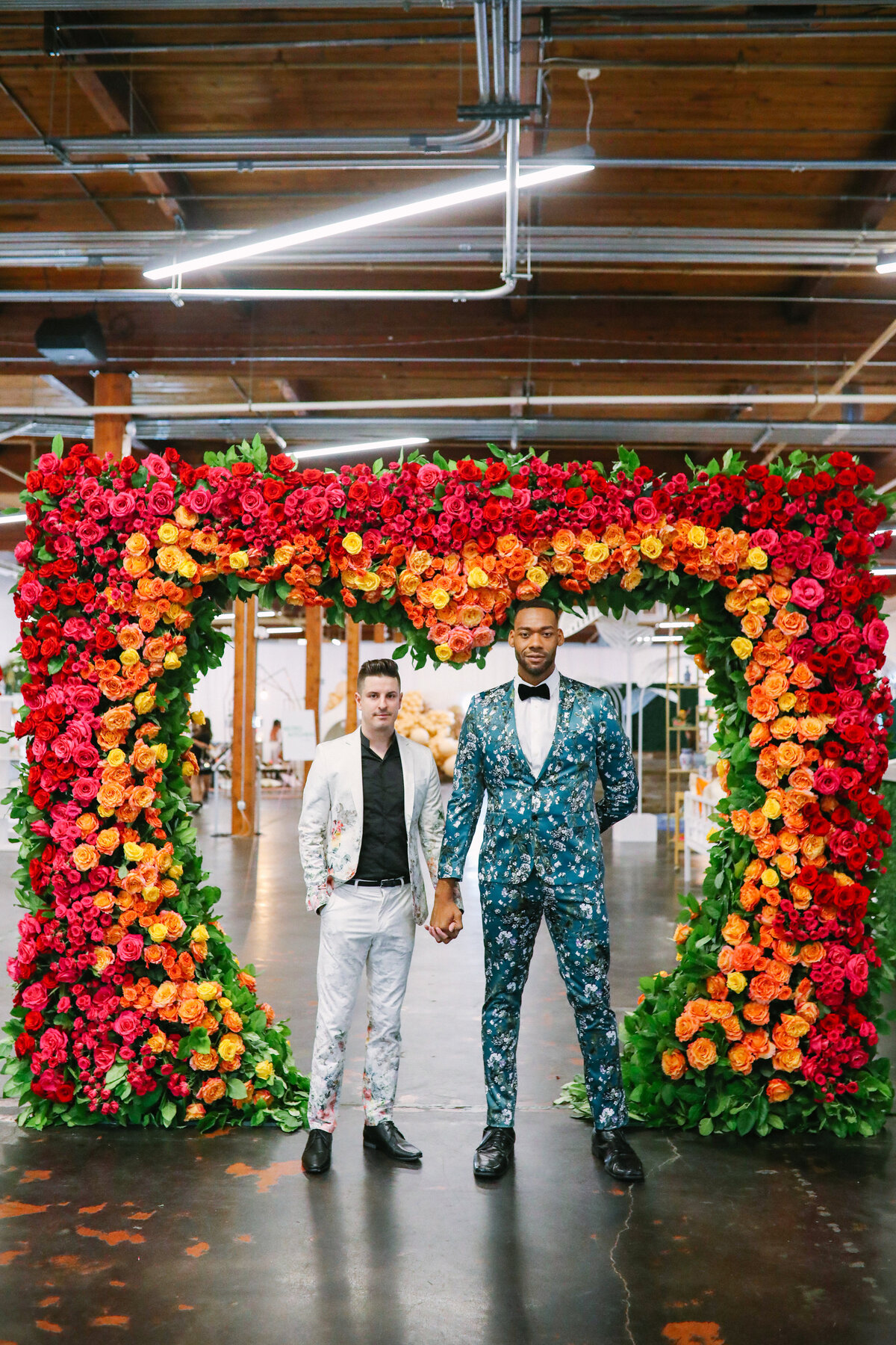Portrait of two grooms posing in front of a large floral arrangement on their wedding day in Dallas, Texas. The floral arrangement is made up of yellow, orange, and red roses to create a large heart shape around the grooms and is backed by greenery. The groom on the right is wearing an elaborate blue suit covered in floral designs and a bowtie. The groom on the left is wearing a white suit with elaborate floral designs and a black dress shirt.