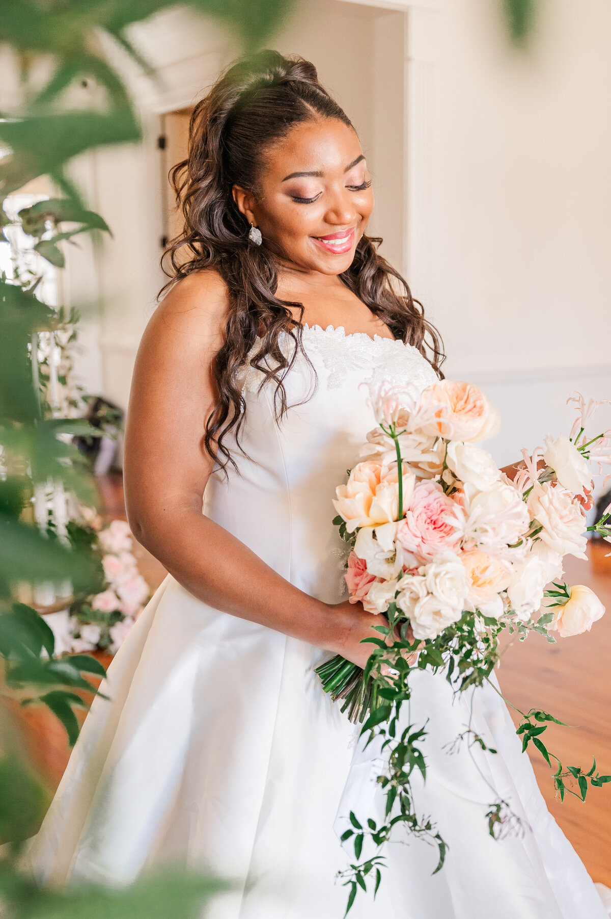 A beautiful African American bride during her bridal portrait session at Donovan Manor while holding pastel pink and white flowers during the springtime as she is surrounded by greenery enjoying her Raleigh wedding photography session with JoLynn Photography
