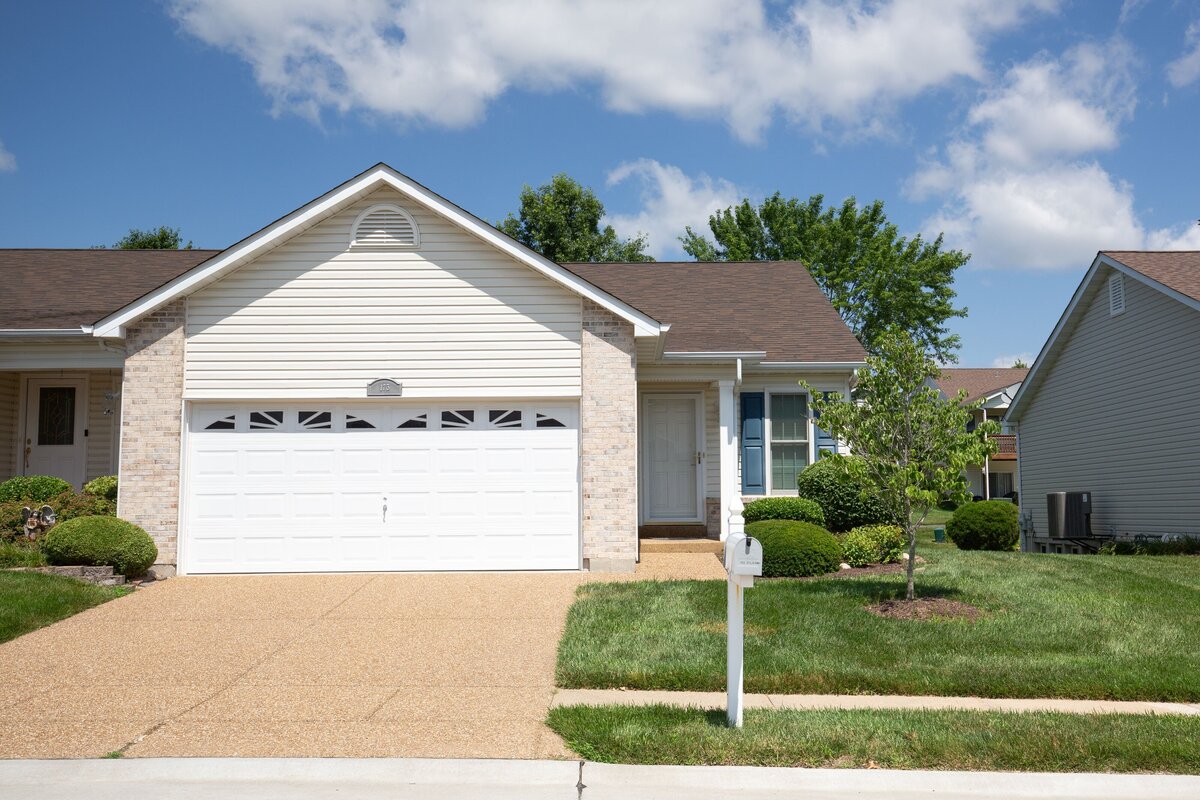 Real estate listing photograph of the front of a cream colored house with light colored brick, and blue shutters. 