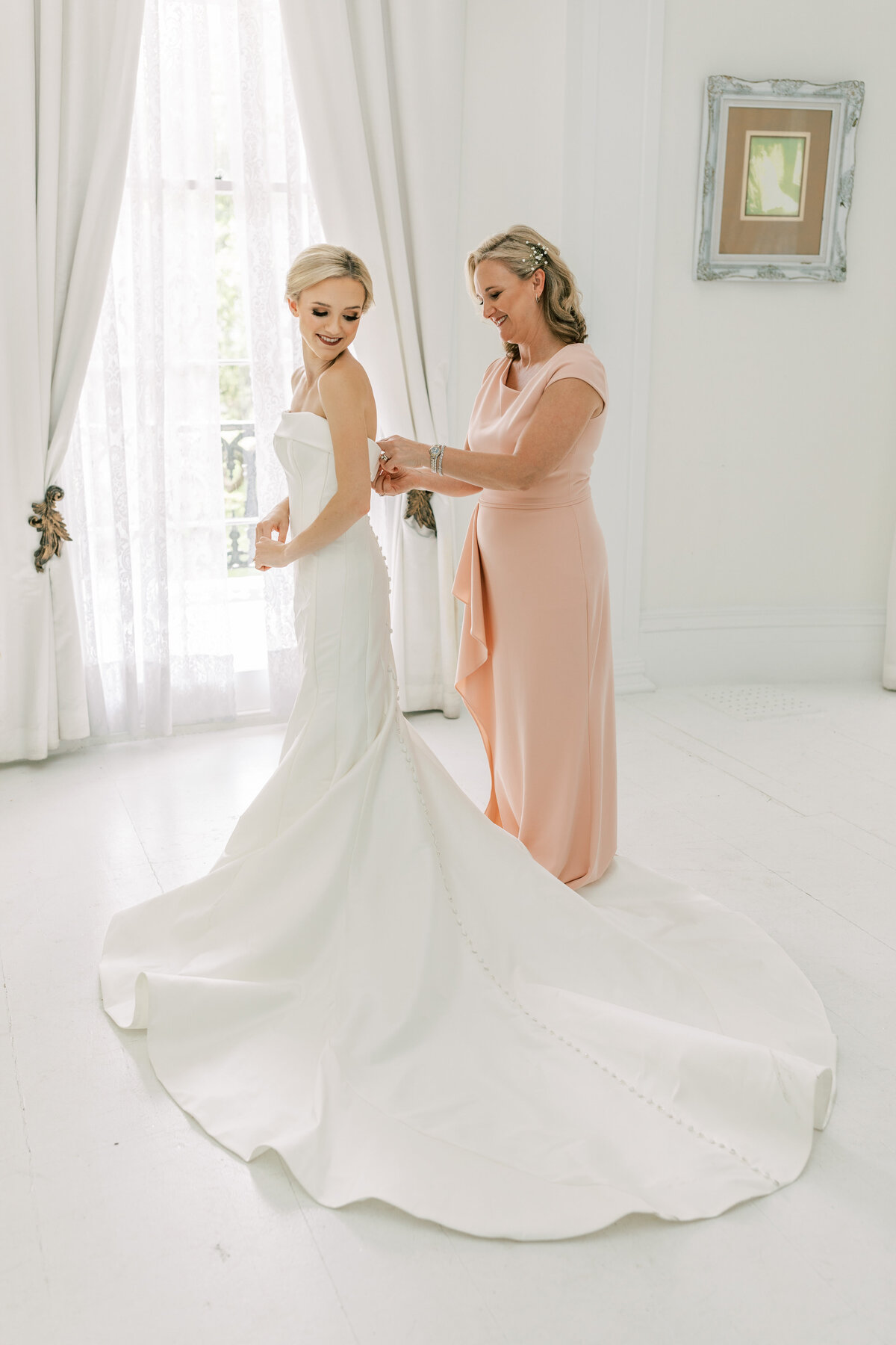 A mom zips the bride up in her dress in a white room.