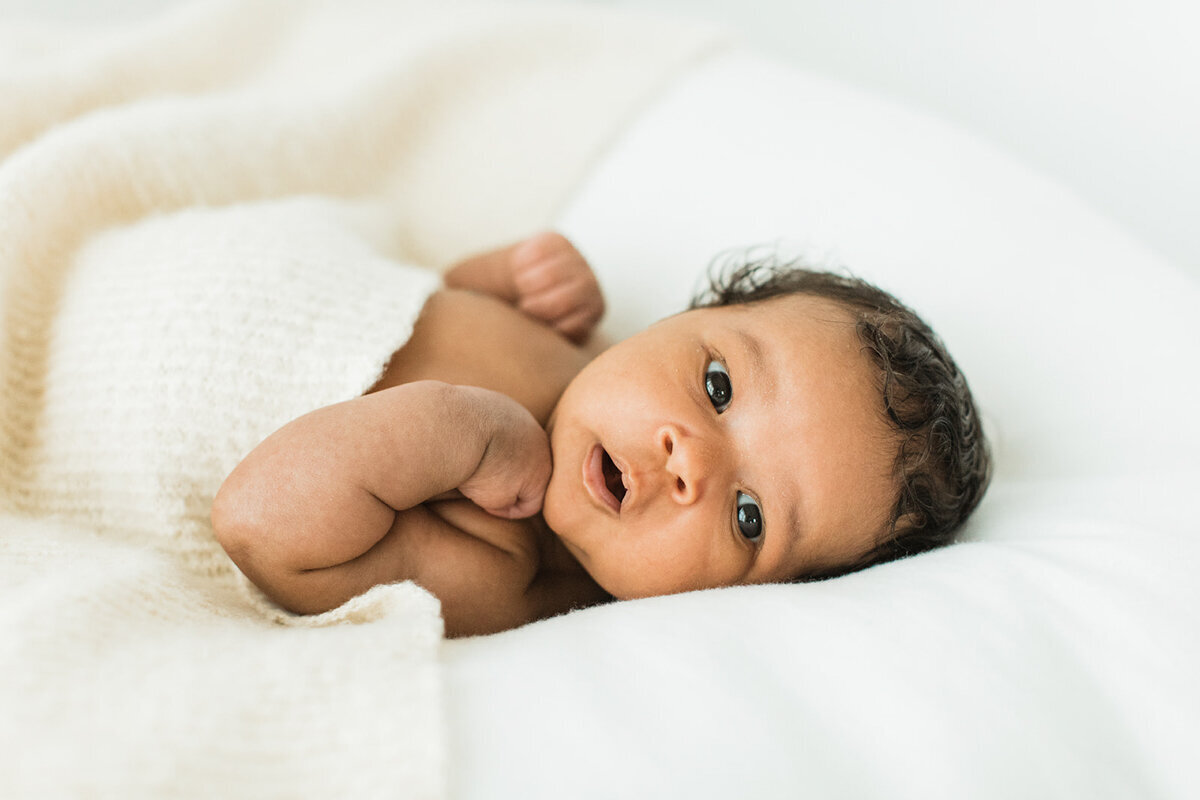 black newborn baby lays surrounded by cozy blankets and looks at camera with cute expression