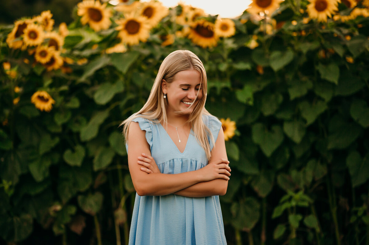 A girl from Waukesha West High School stands in a Hartland, WI field of sunflowers wearing a denim sundress and looking at the ground in front of her.