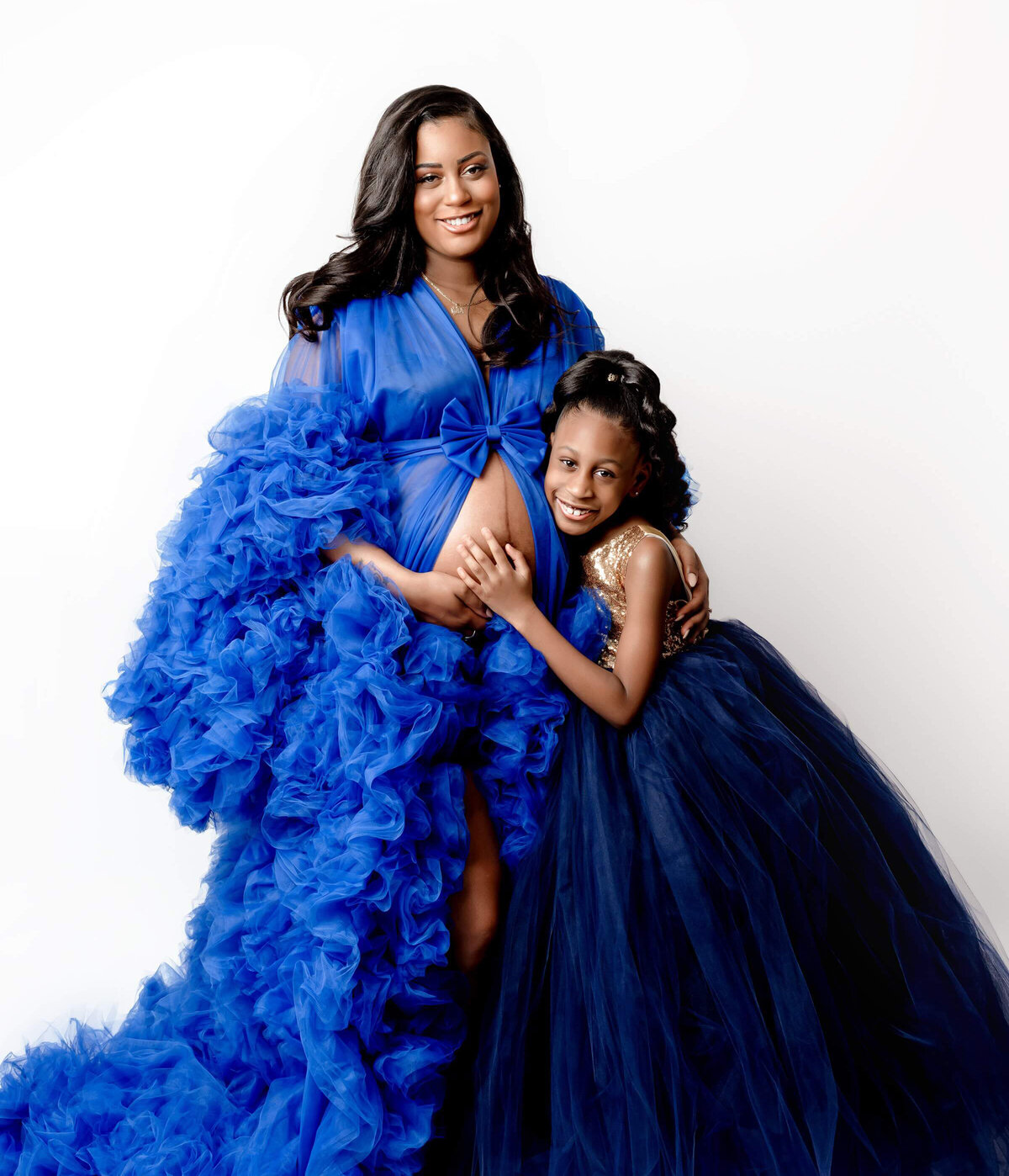 A mother and daughter pose together in a maternity studio photo shoot. The mother, with her baby bump proudly displayed, lovingly embraces her daughter.