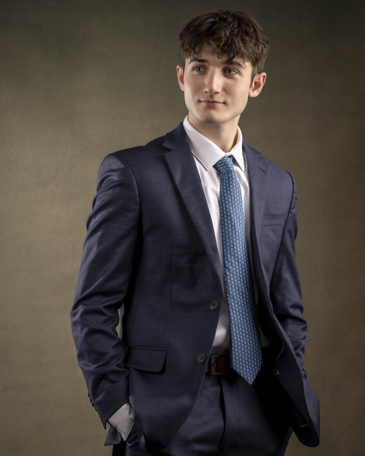 Fine-art style headshot in CT of a young man with short brown hair wearing a blue suit, posed with relaxed confidence against a dark studio background, by Rob Faber Photography, expert in headshots in CT and MA.
