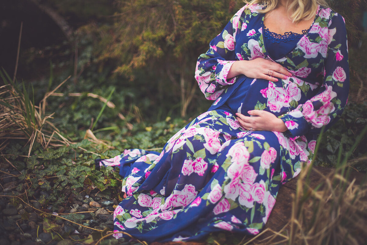 An expecting mom in a bright floral dress sitting on the forest floor.