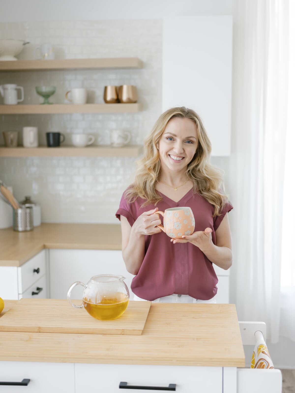 Woman wearing a pink top holding a coffee mug while making tea in a kitchen and smiling at the camera