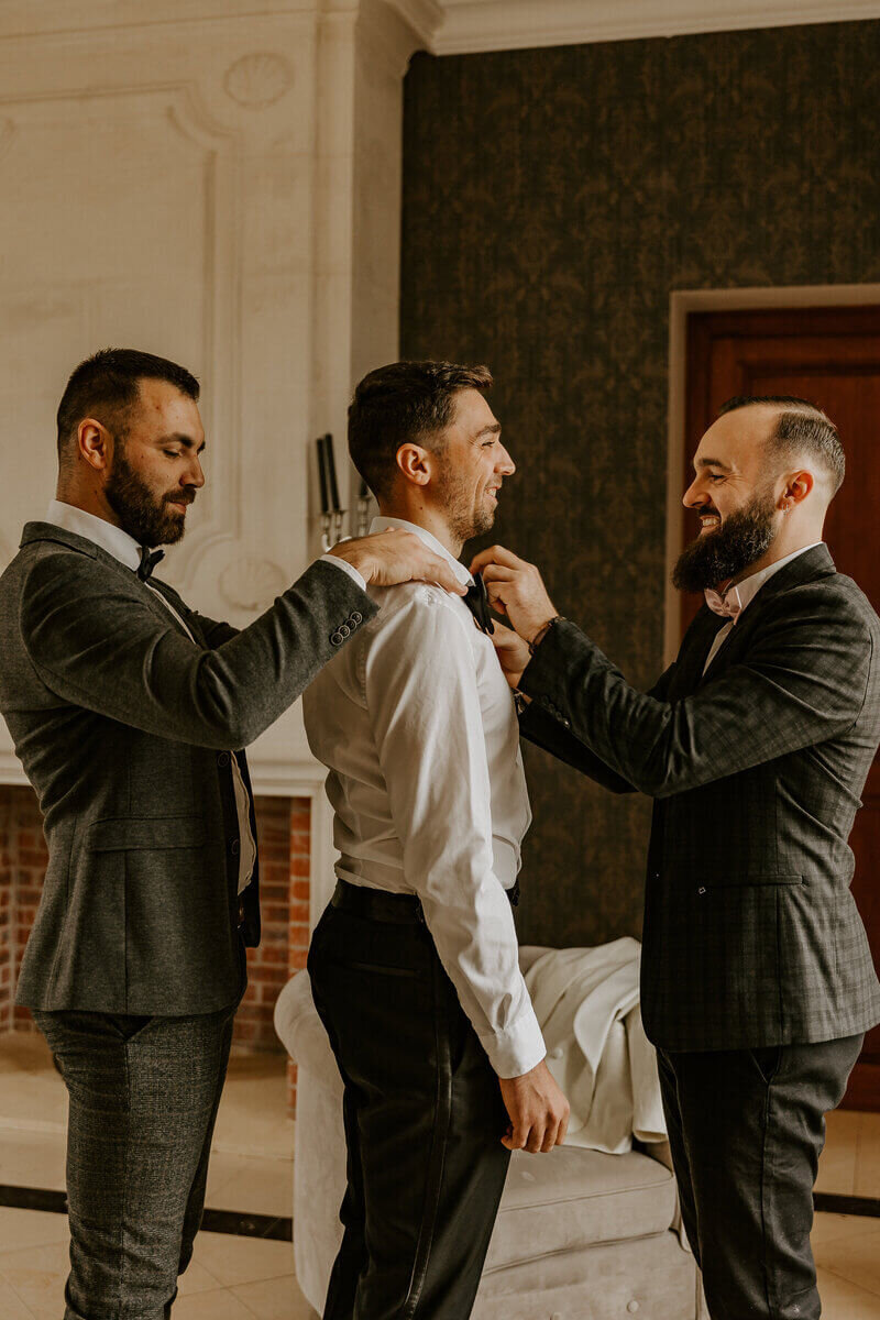 Deux témoins ajustant le noeud papillon du marié dans une pièce décorée d'un fauteuil et d'une vieille cheminée. Photo issue du workshop photographie de mariage.