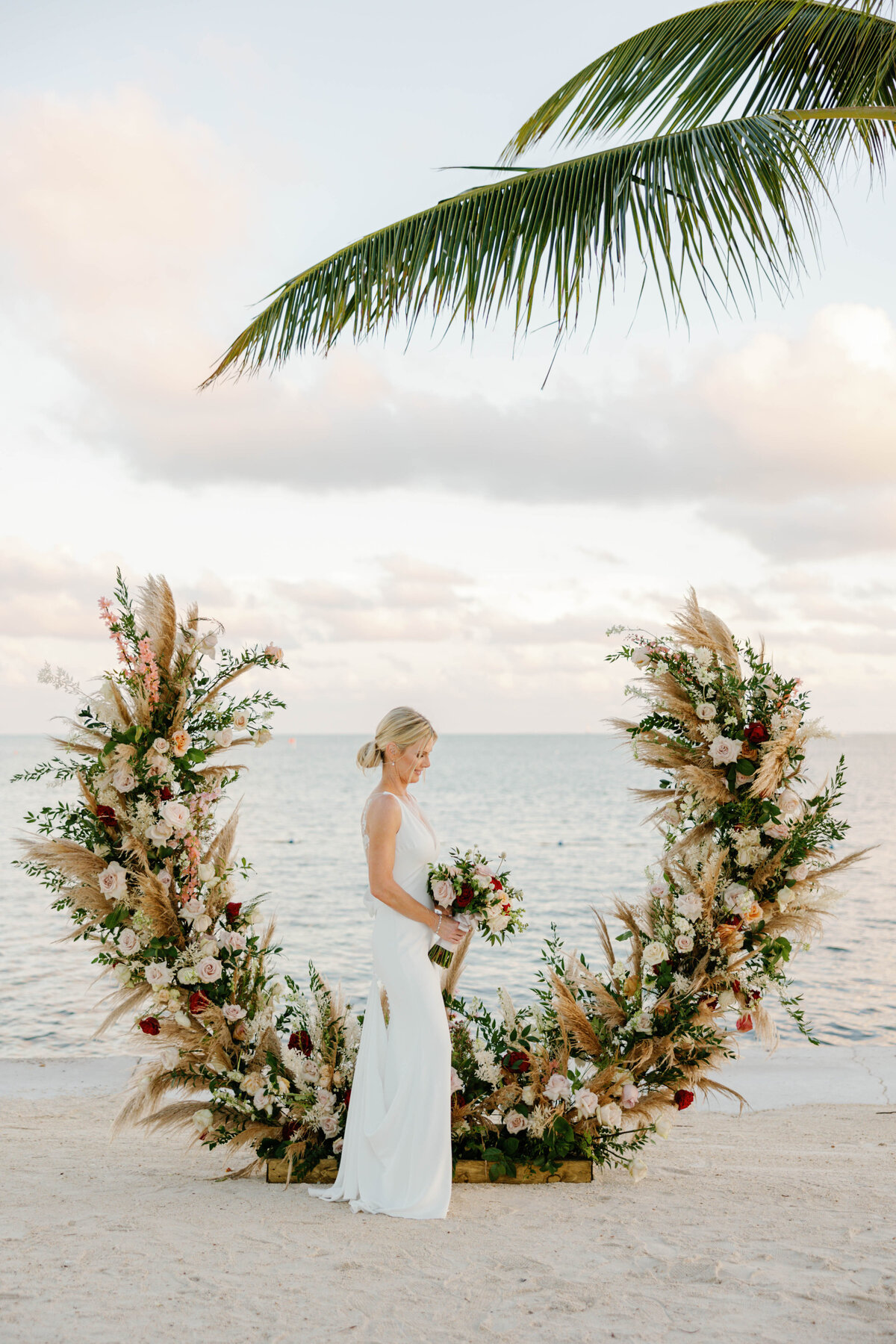 Bride standing in front of a floral arch on the beach, captured by Claudia Amalia Photography in Miami, Florida.