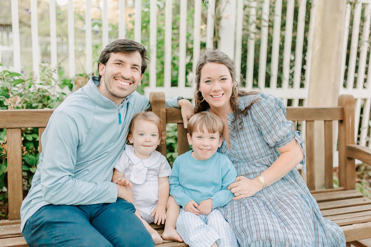 Joyful outdoor family portrait—parents and their two young children sit on a wooden bench, dressed in coordinated blue tones. Capturing authentic moments with a Birmingham, AL family photographer
