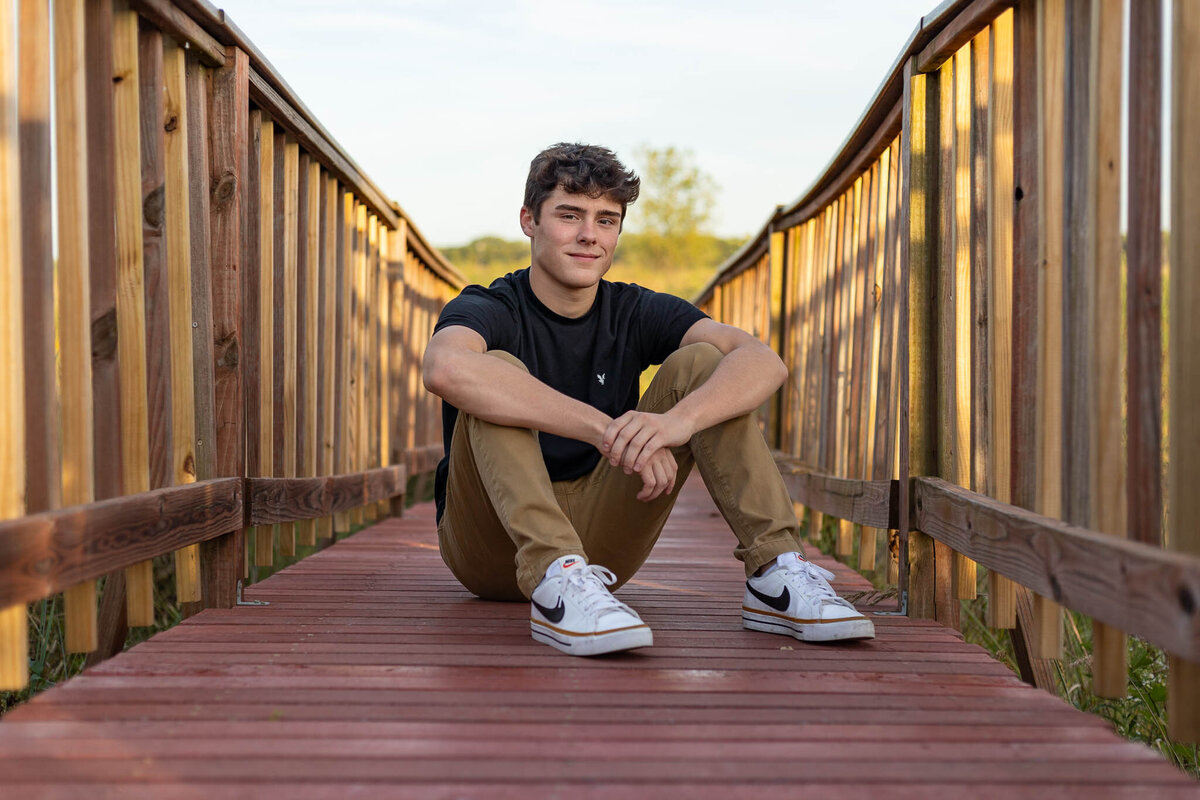 Capture the essence of senior year with a bold and natural outdoor portrait. This stylish senior session features a young man seated on a rustic wooden bridge, bathed in golden hour light. Megan Bowers Photography specializes in authentic, expressive, and timeless senior portraits. Book your session today!