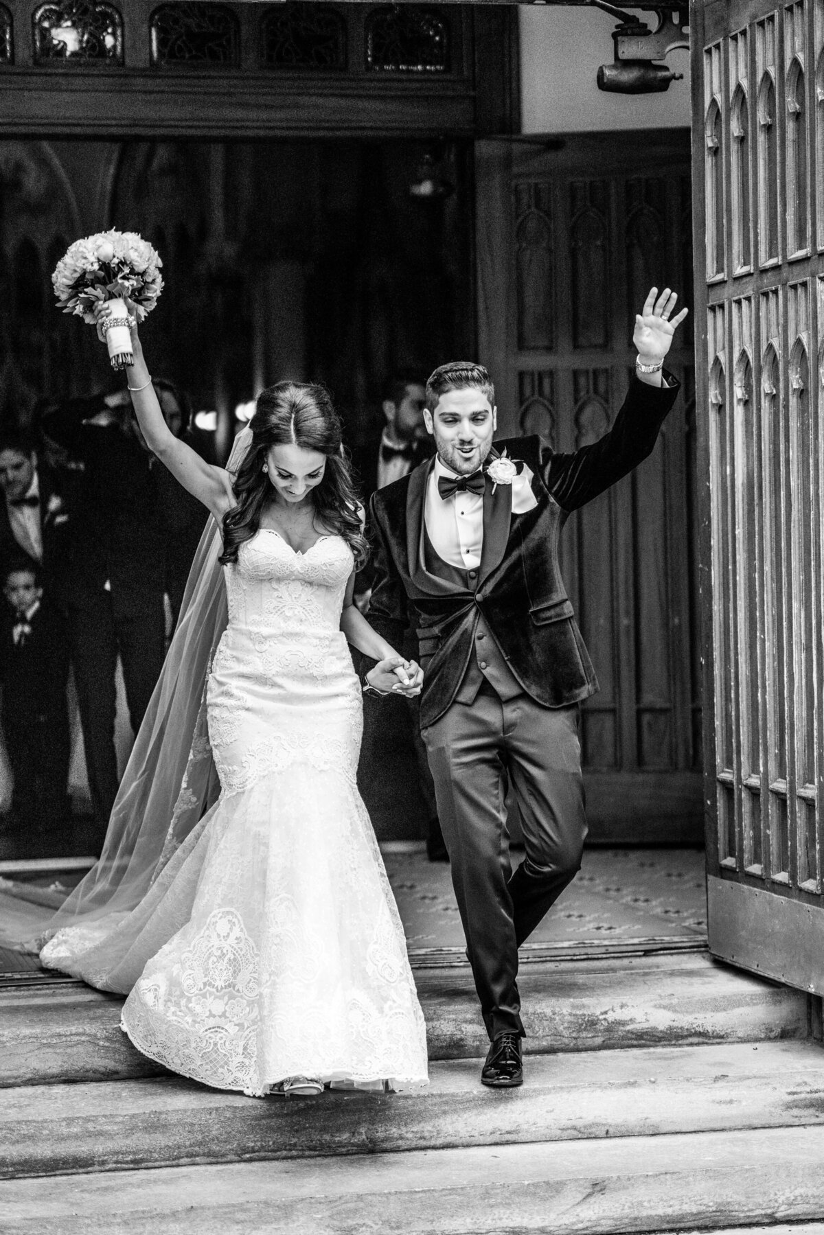 In this black and white image, the couple exits the church, celebrating their marriage with joyous expressions. The timeless quality of the photo captures the excitement and emotion of their post-ceremony moment, highlighting the significance and happiness of their wedding day.