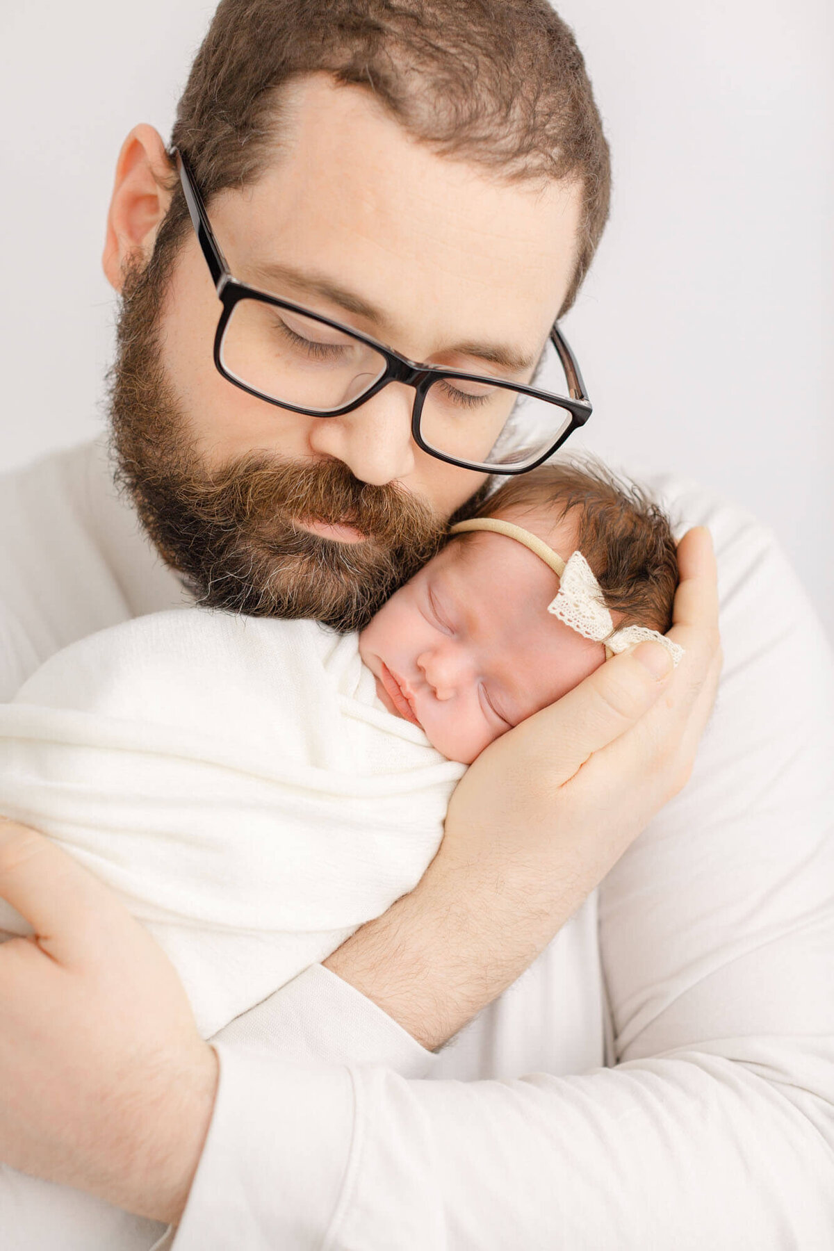 Dad dressed in a white shirt holding baby up to his face, they are cheek-to-cheek. Dad is wearing black glasses and his eyes are closed. Baby is sleeping peacefully.