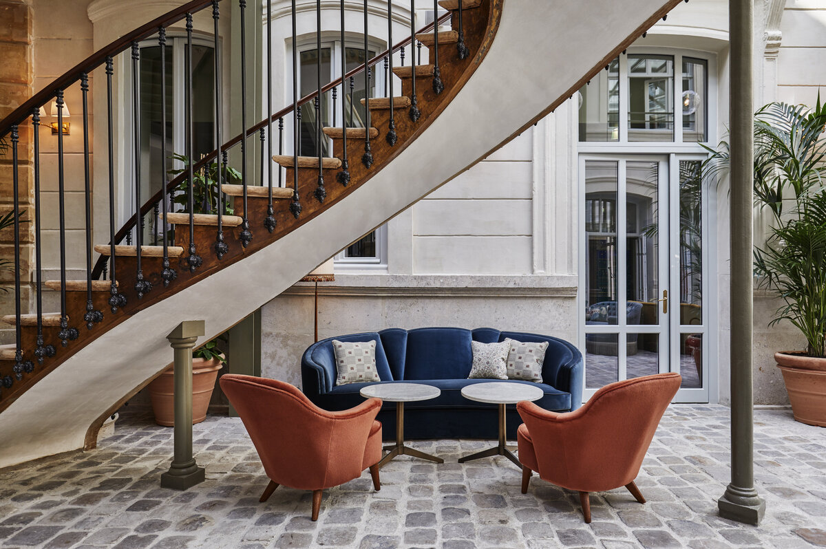Antique Parisian Staircase above seating area with blue velvet curved sofa, two marble cocktail tables and red velvet vintage armchairs