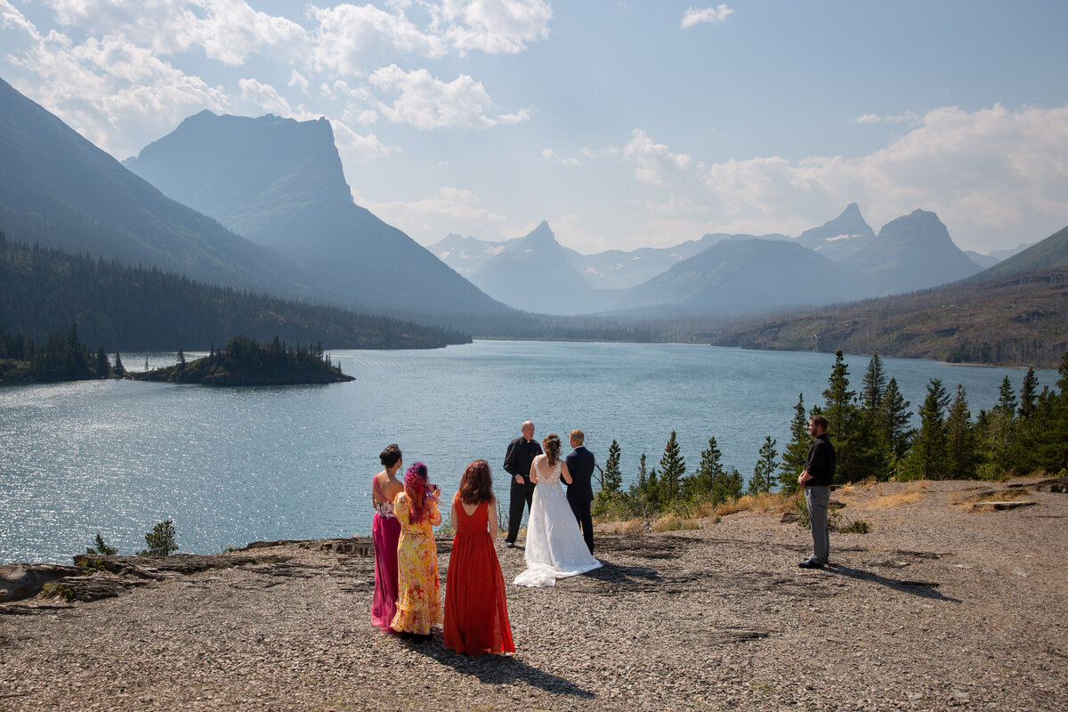 A group stands on top of a point overlooking a lake and snow capped mountains as the bride and groom have their elopement ceremony.