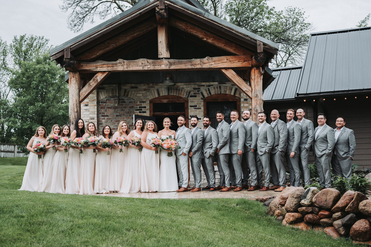 Full wedding party standing in front of a wood and stone bridal suite outdoors.