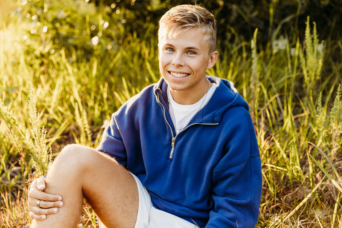 blonde senior boy in a blue sweater sitting in the grass