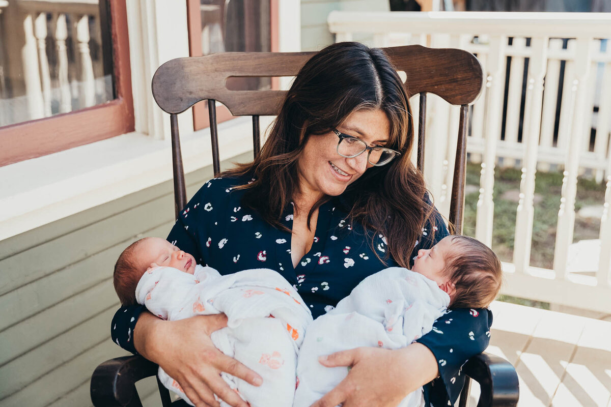 a mother holds her twin newborn babies while sitting in a rocking chair on her home's front porch