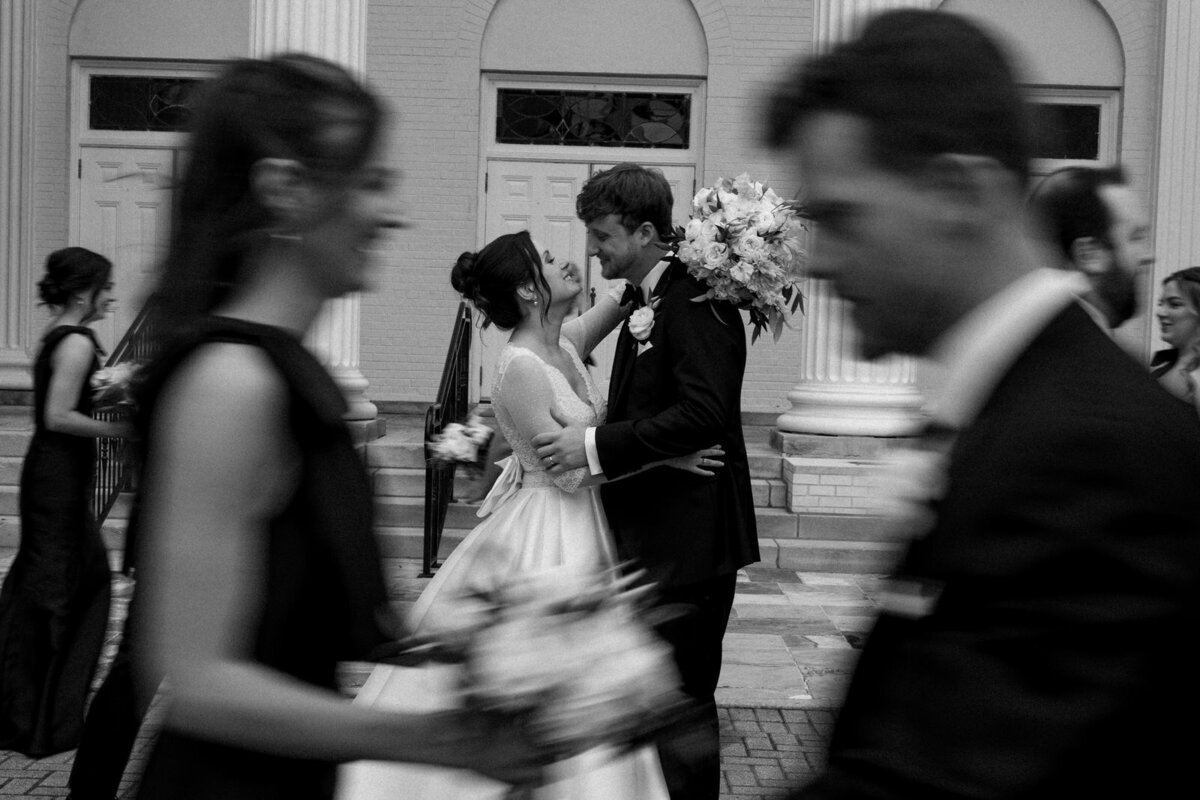 Bride and Groom kissing while wedding party laughs and celebrates around them, captured by Andrea-Marie Photography