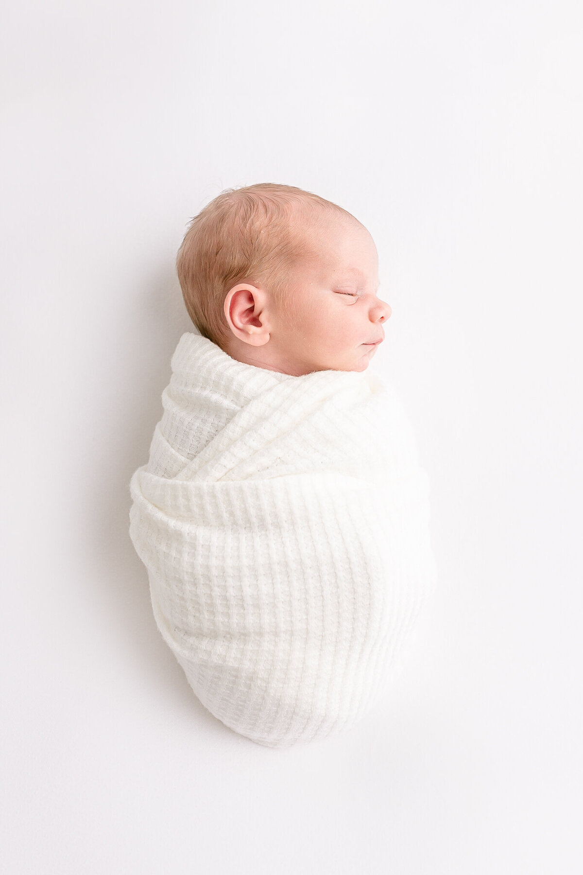 Sleeping baby swaddled in white laying on white backdrop. All swaddled and sleeping peacefully on back with head turned to one side. Image taken during PDX newborn photography session.