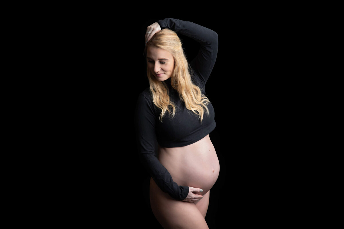 Blonde pregnant woman wearing a black crop top standing in front of a black backdrop with her arm up for her studio maternity photography session in Burlington, ON