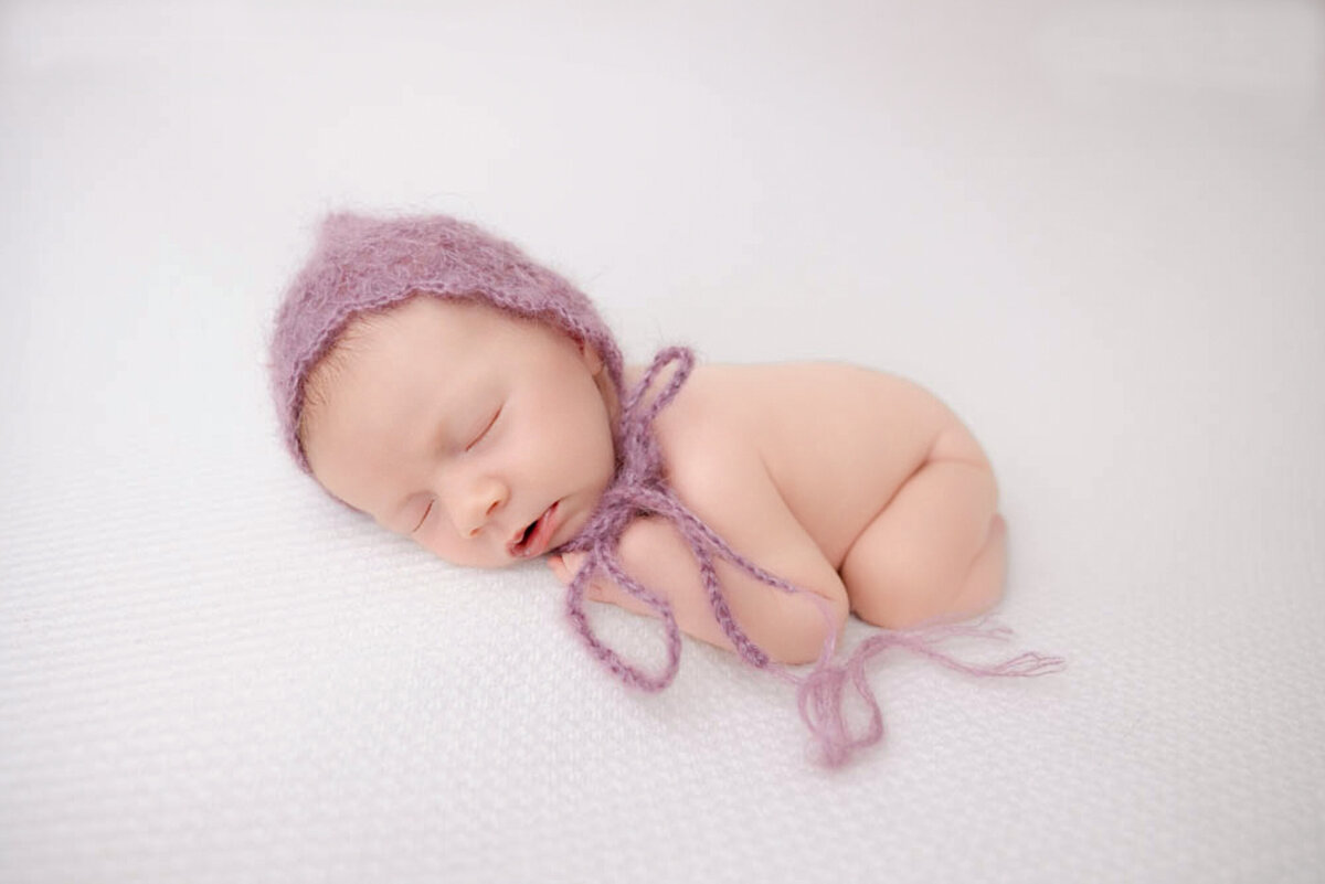 two-week baby girl posed lying on white blanket and wearing a purple bonnet at Heather Nicole Photography.