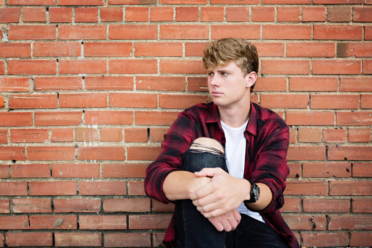 Guy in flannel shirt and torn jeans sits against a brick wall