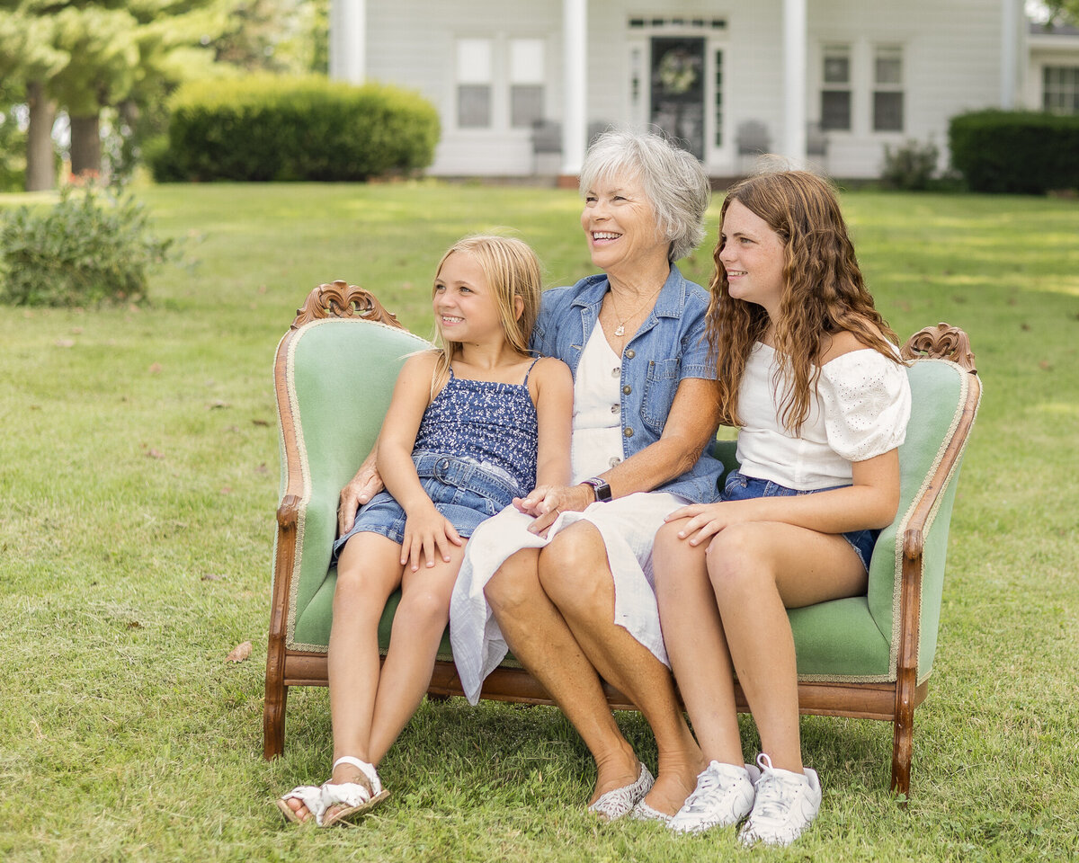 Sisters and grandma are sitting on a vintage green couch in the grass with their family two story home in the background, looking away from the camera smiling. Older daughter is wearing blue jean shorts, a white short sleeve shirt, and has long brown hair.  Youngest daughter has a dark blue and white tank top style shirt with a blue jean skirt, and has medium length blonde hair.Grandma is wearing a white button up dress with a jean jacket over it, with short white hair.  