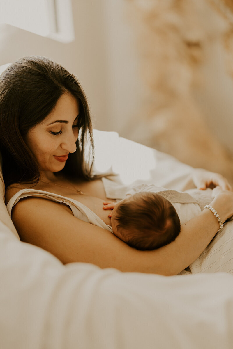 Maman souriante, donnant le sein à son bébé dans un décor cosy, posant pour Laura lors d'une séance photo naissance.