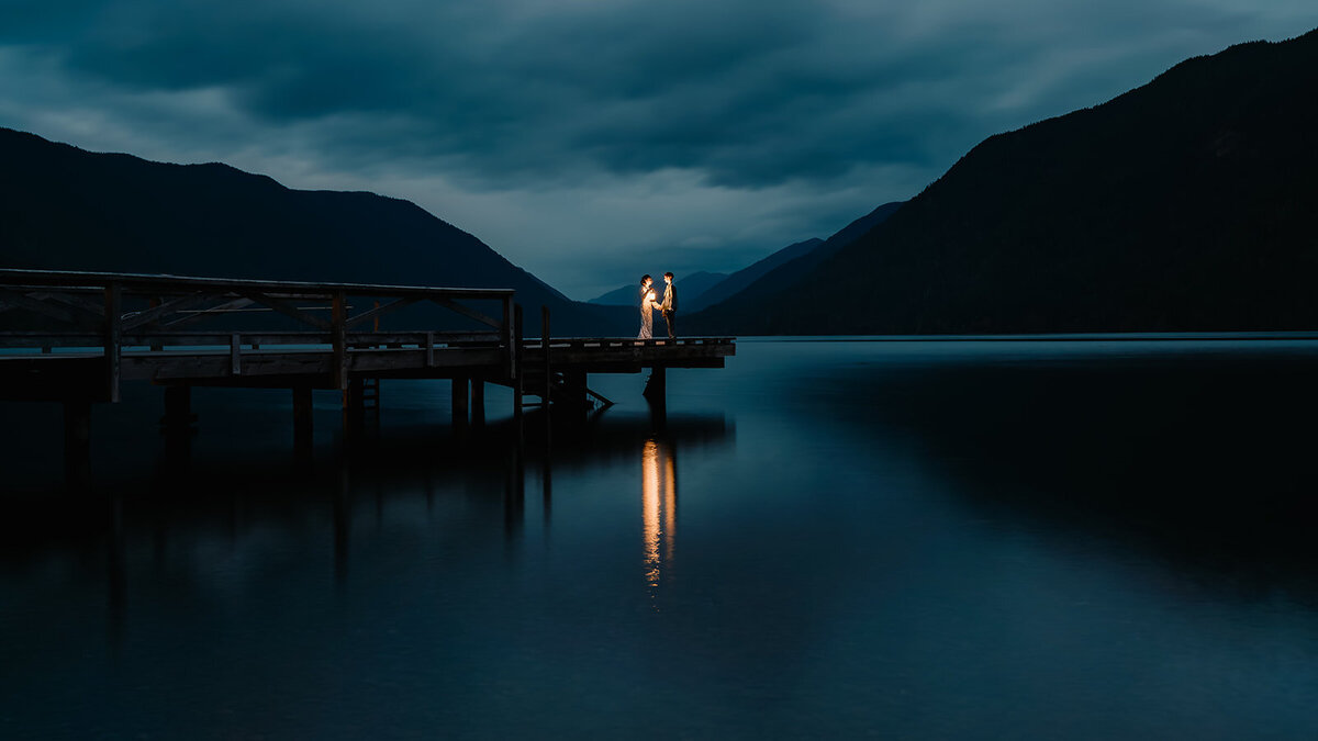 taken just after sunset two brides in their wedding attire stand at the edge of a dock over a deep navy lake. They hold lanterns as they embrace. Their adventure elopement photographer takes a long exposure shot making the water look like glass, framing them between the mountains, and catching the reflection of their lanterns in the water.