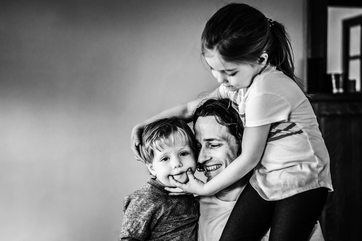 An older sister sits on her father's shoulder , as she uses her hands to make her little brother smile during an in home photoshoot.