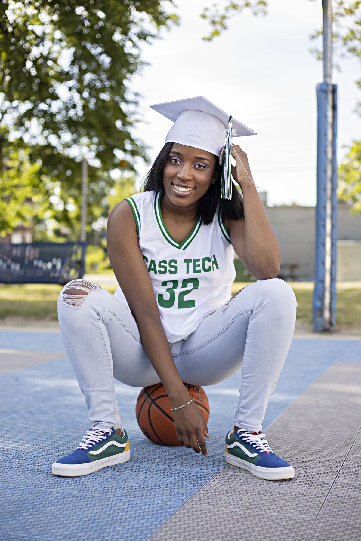 Senior girl with Cass Tech basketball jersey and white graduation cap sitting on basketball at basketball court