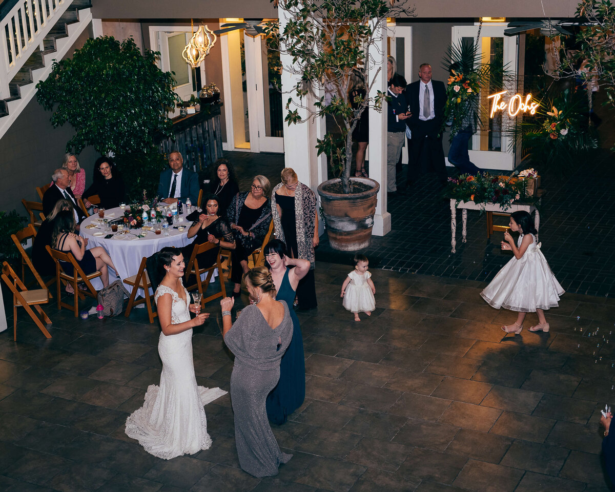 A bride in a white gown dances with a woman in a gray dress at a wedding reception, while guests seated at round tables watch and smile. 