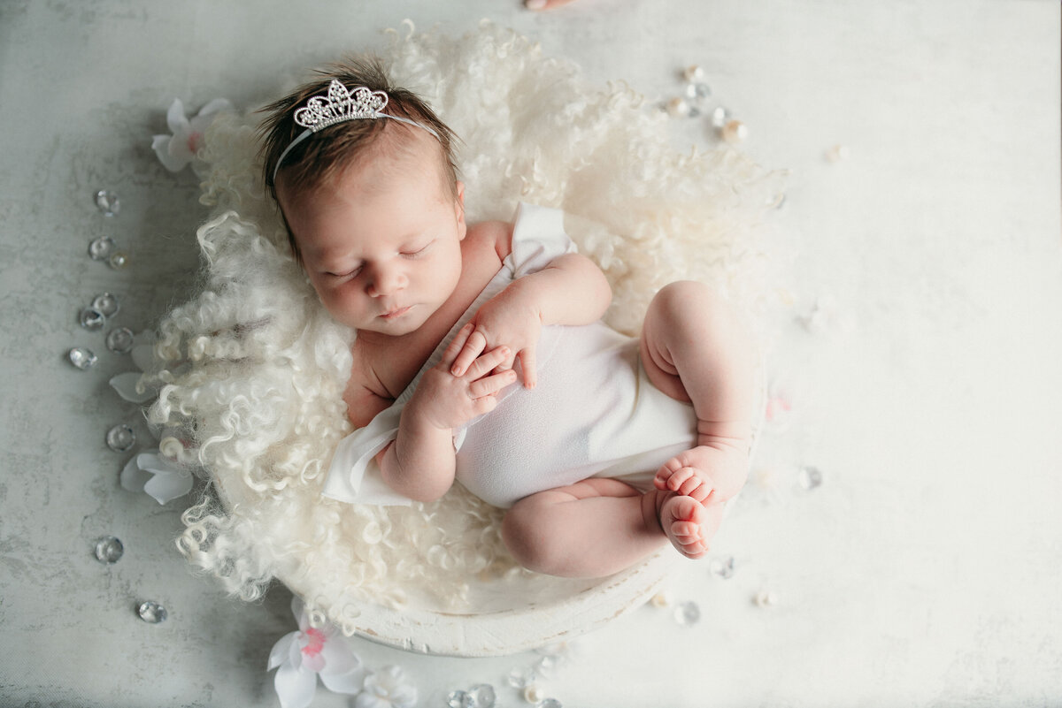 Newborn girl in white outfit wearing a tiara laying on white fur on a white backdrop