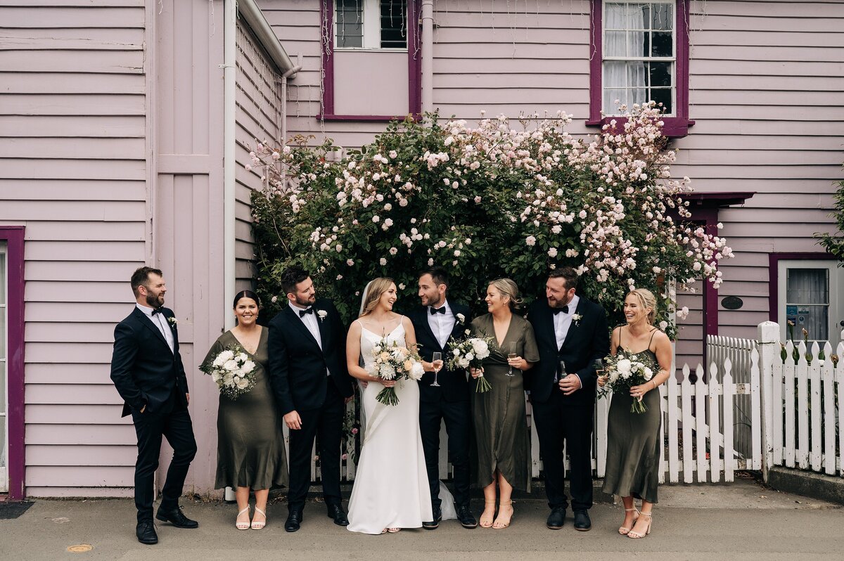 bridal party with olive green dresses and black suits in front of pink house in akaroa wedding new zealand