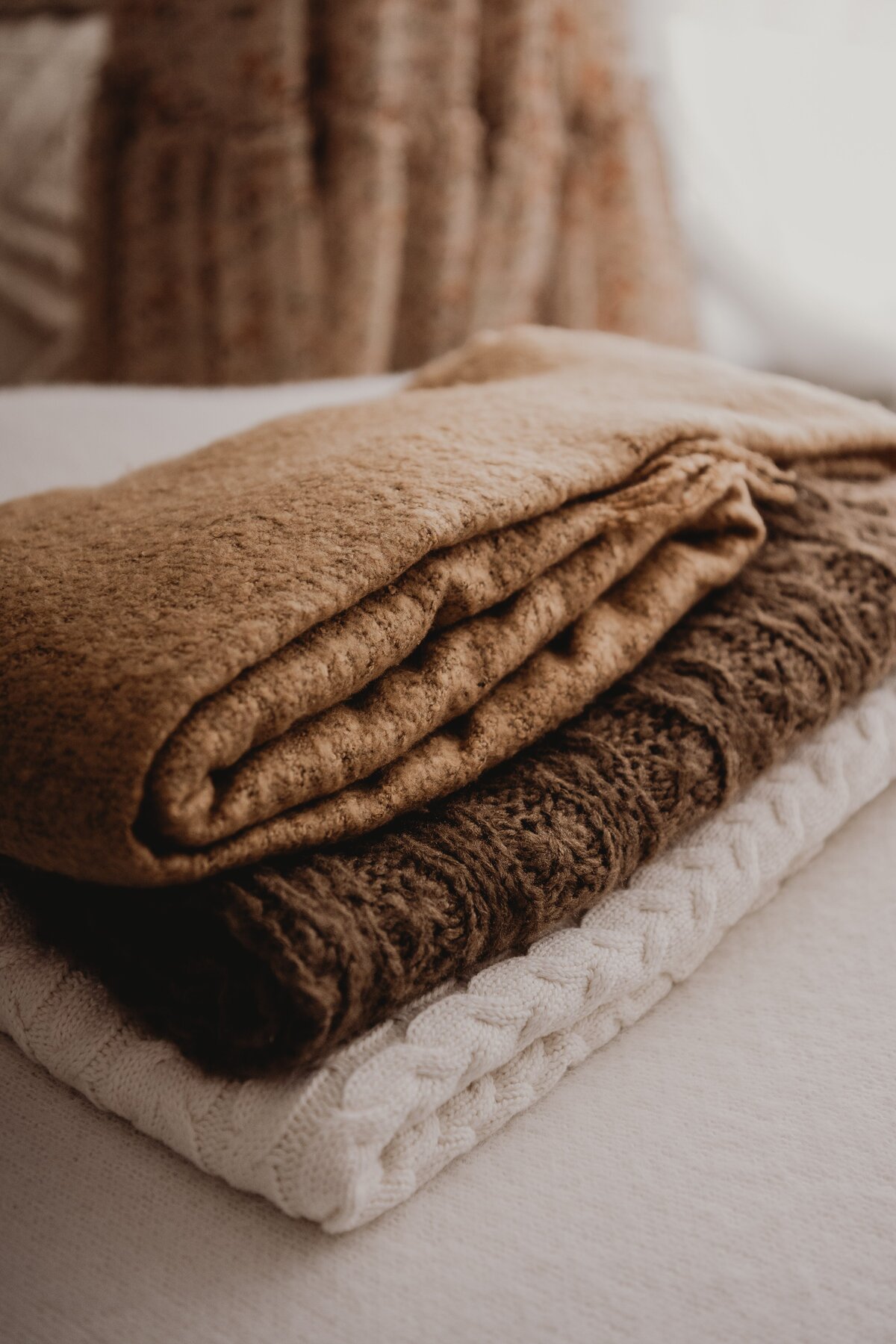 Three neatly folded blankets—one brown, one dark brown, and one white—stacked on top of each other on a light-colored surface, evoking the cozy warmth often captured in Wollongong motherhood sessions.