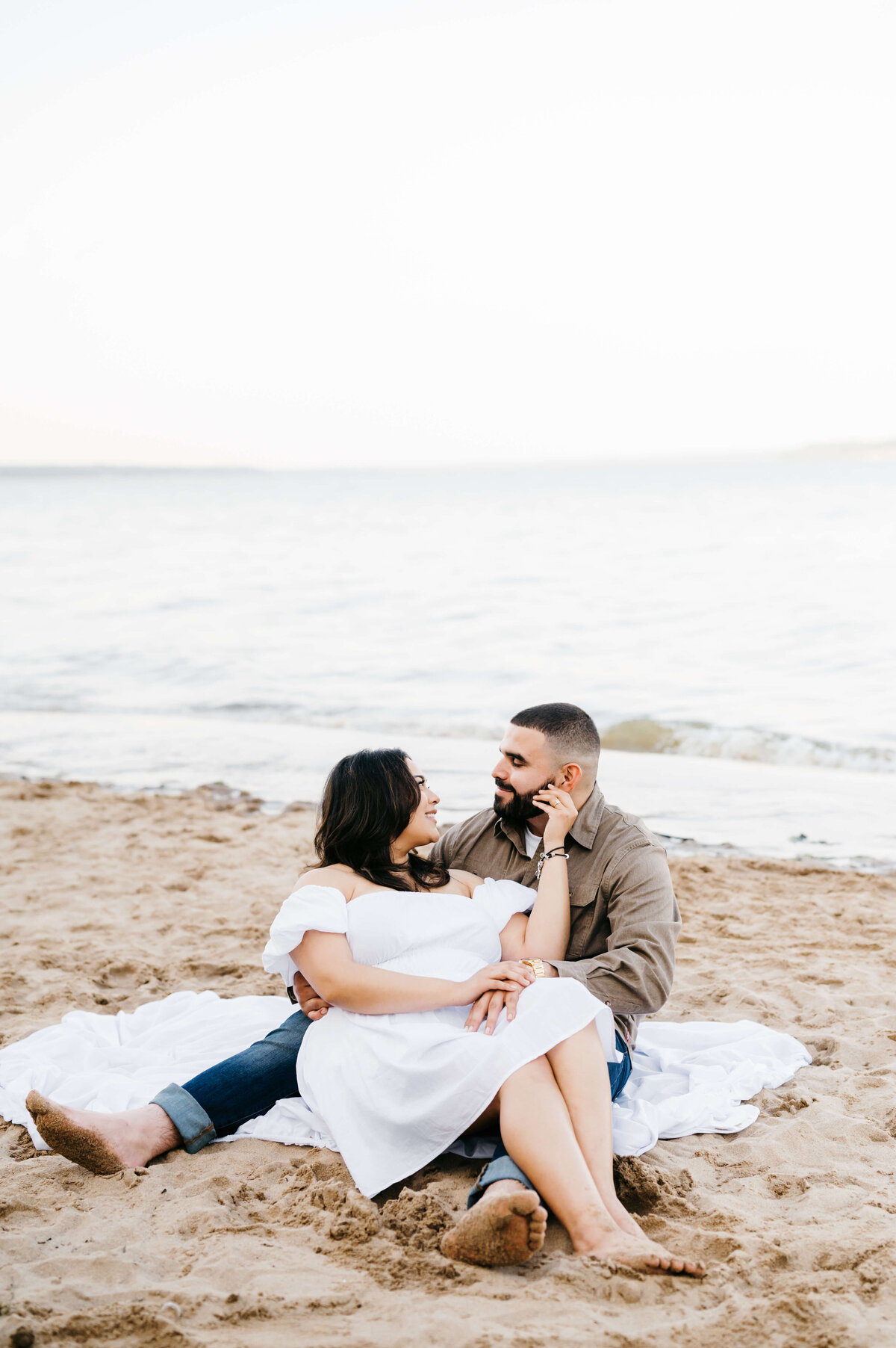 Virginia wedding photographer captures beach engagement photos with man and woman sitting on a white blanket together with the waves coming in behind them