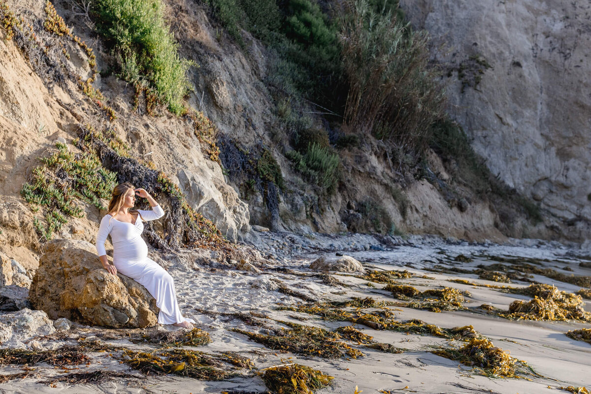A pregnant woman in a long white dress sits on a rock at a beach. Shes gazing into the distance, with one hand on her belly and the other shielding her eyes. The background features rocky cliffs and scattered seaweed on the sand.