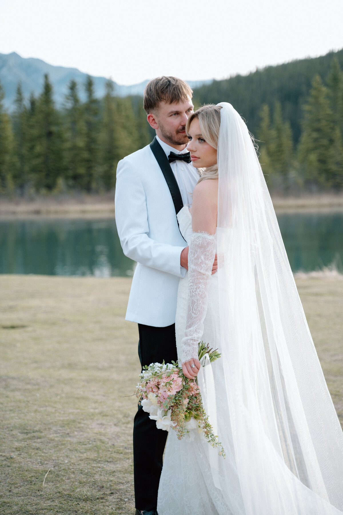 Groom lifts bride’s veil in scenic lakeside meadow with Banff mountains behind.