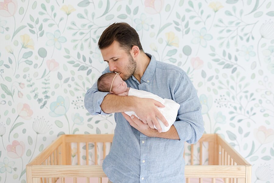 dad holding baby and kissing her in front of crib and wallpaper accent wall by DC Newborn Photographer Emily