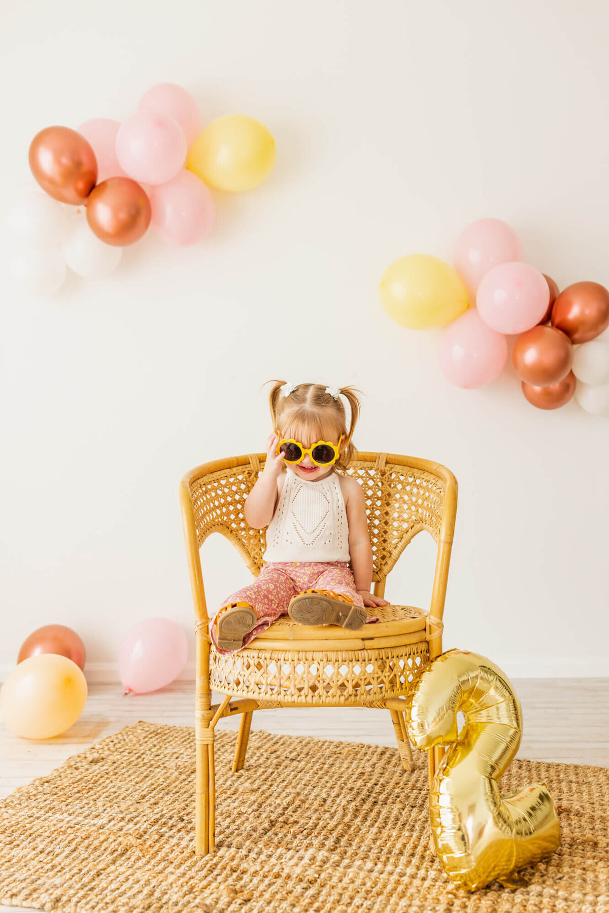 Toddler girl dresses in bell bottoms and a lace romper sitting on wicker chair with sunglasses on