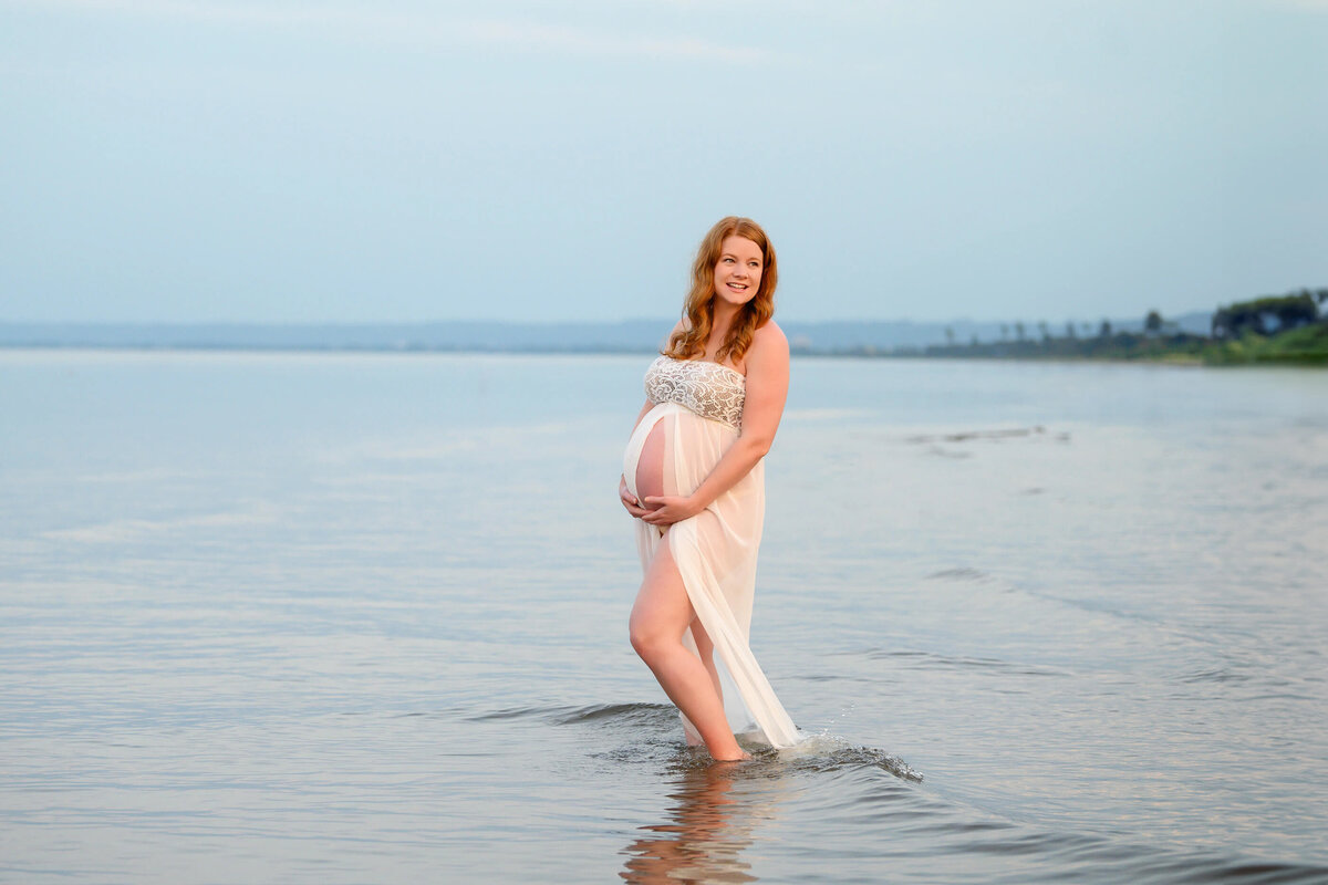 pregnant woman wearing a white maternity dress standing in the water for her sunset beach maternity photos in Burlington, On