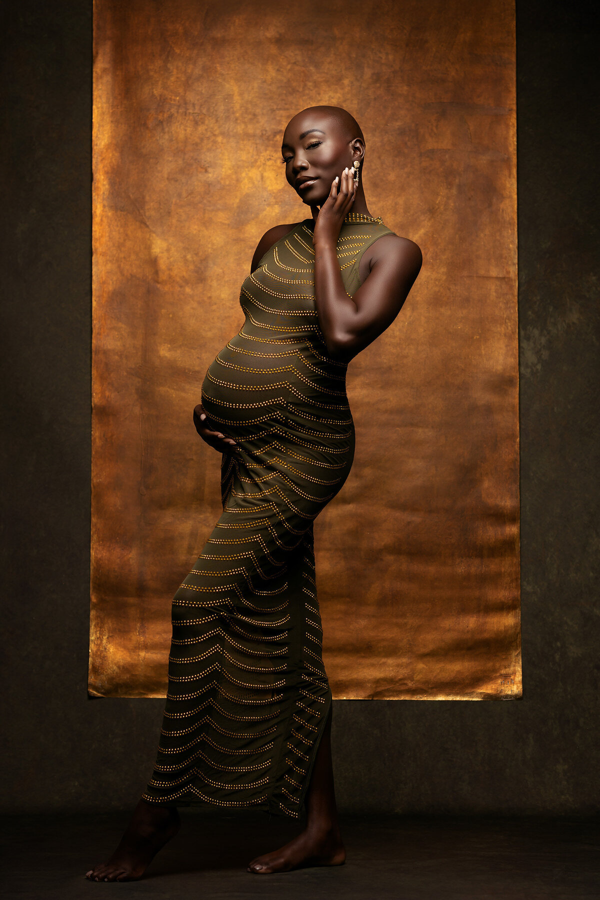 A beautiful expecting mother dressed in an olive-green gown adorned with golden detailing, standing gracefully against a rich, golden backdrop. Her serene expression and poised hand enhance the elegance of this Dallas maternity photography session.