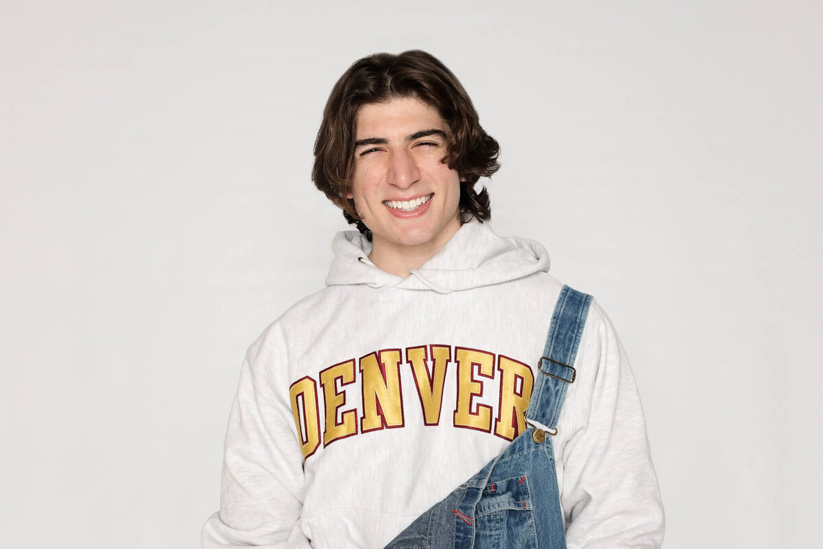 A person with medium-length dark hair smiles at the camera, wearing a white hoodie with DENVER written on it and denim overalls over one shoulder. The background is plain and light-colored.