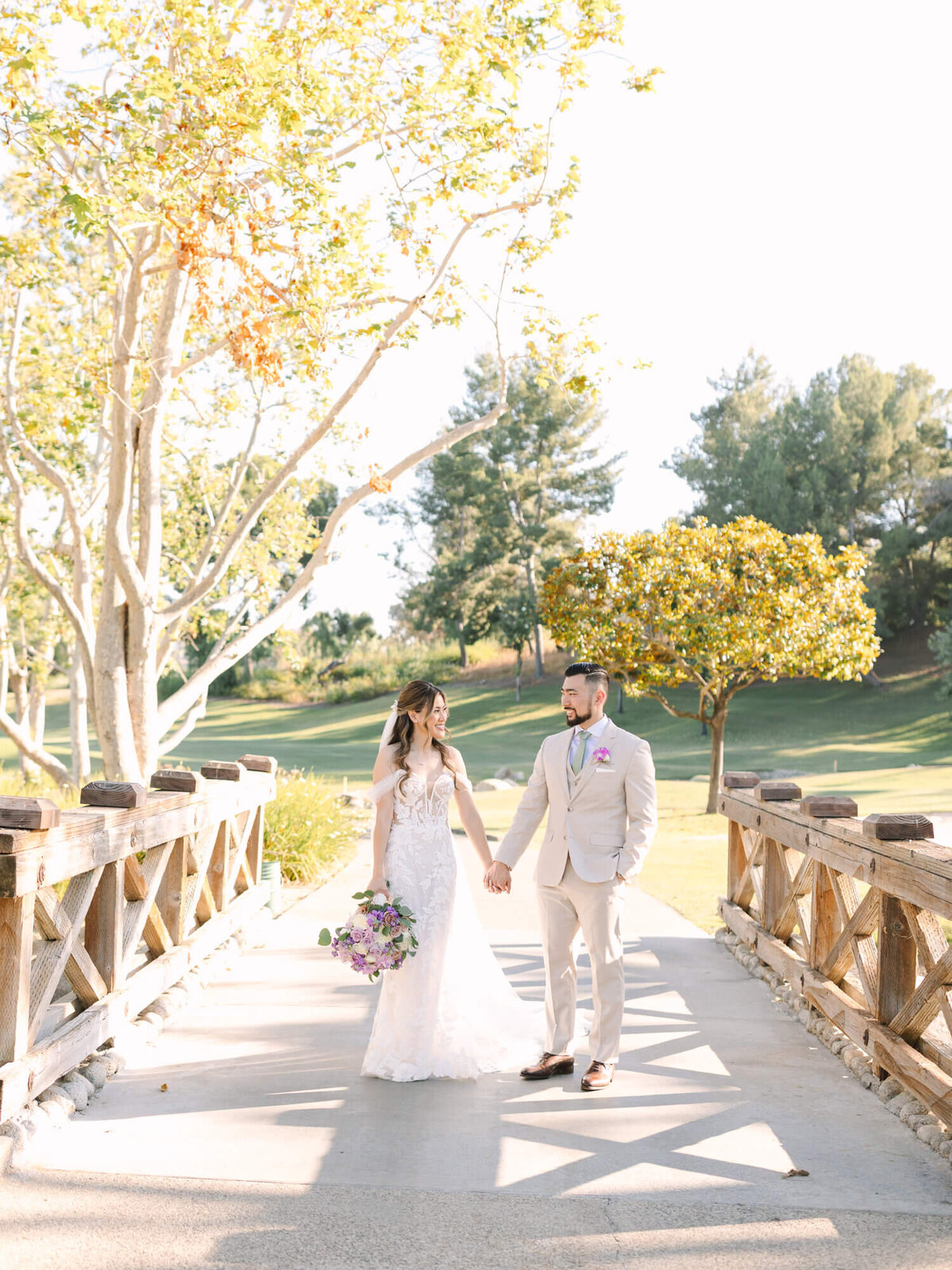Bride in lace gown holding a bouquet and groom in beige suit walk hand in hand on a sunlit bridge, surrounded by green trees and a bright sky.
