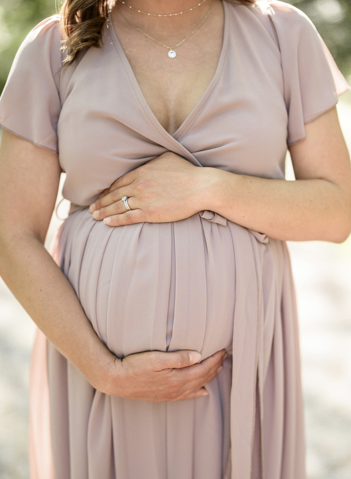 Close-up of a woman cradling her belly in a soft mauve dress during a sunlit maternity session.