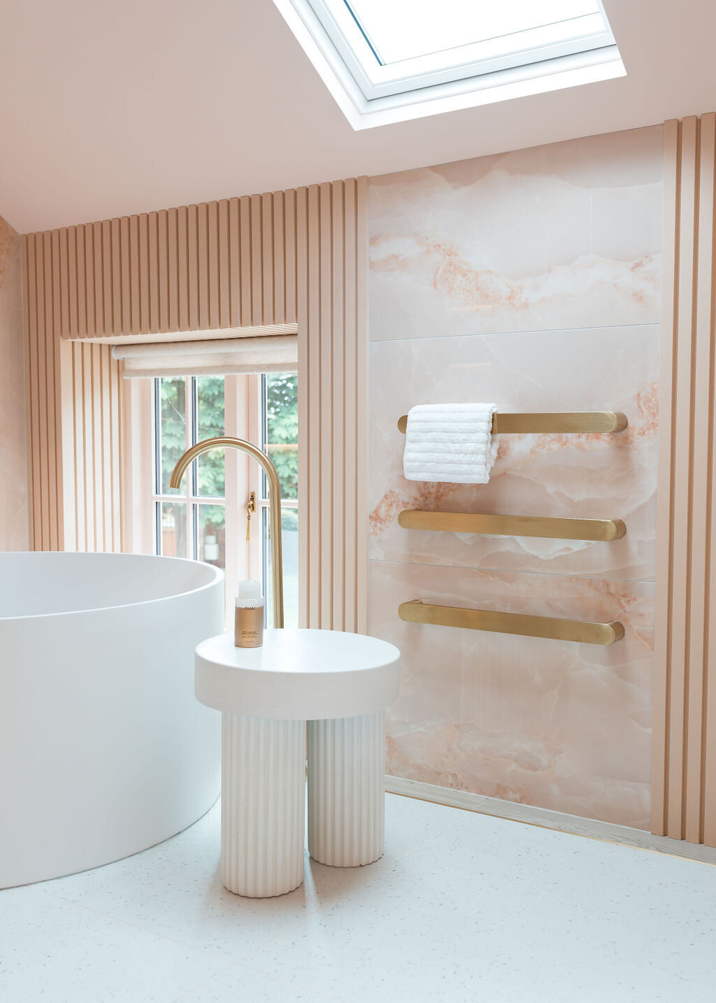 A modern bathroom featuring a round white bathtub with a sleek gold faucet. A small white table stands next to the tub. Three white towels hang on elegant gold bars mounted on a pink marble wall. A window with vertical blinds adds natural light.