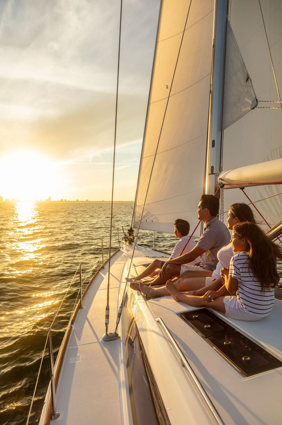 A family of four relaxes on the deck of a sailboat, enjoying a sunset over the water. The sky is clear, and the sun casts a golden glow on the sails and sea.